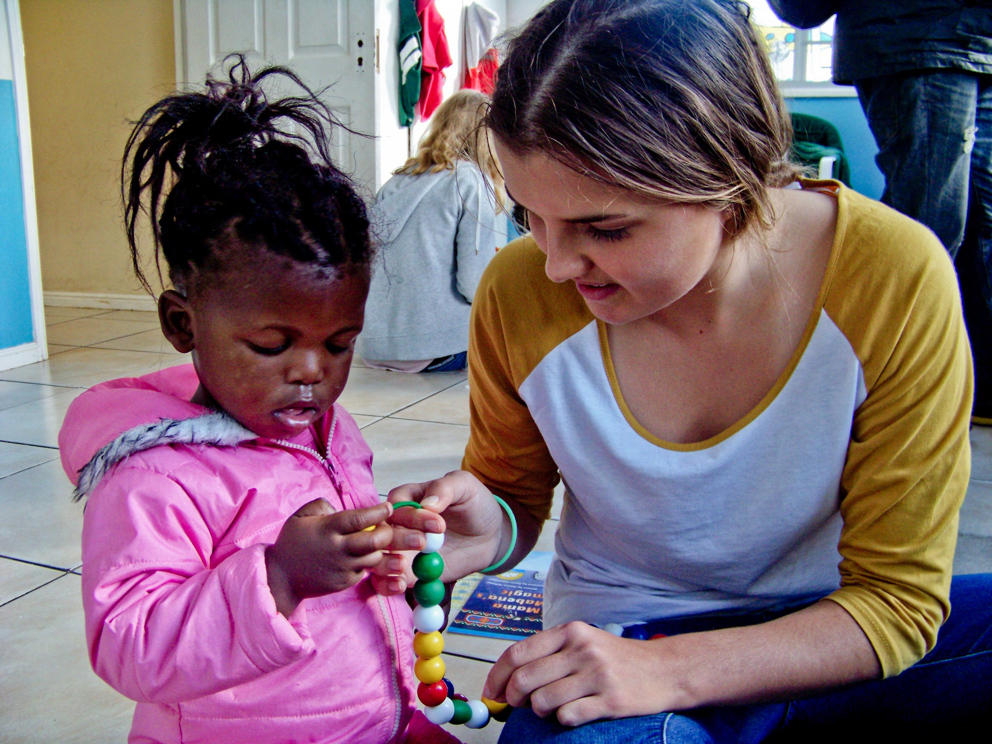 Female volunteer with children in South Africa helps young girl during a craft activity at a Childcare placement. Projects Abroad Childcare volunteer helps a young child withs crafts in South Africa.